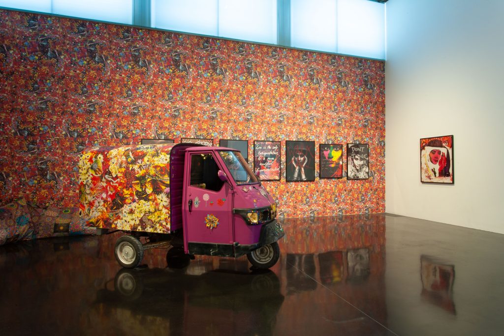 A decorated tuk tuk in front of a colourful wallpaper background and artworks by Chila Kumari Singh Burman